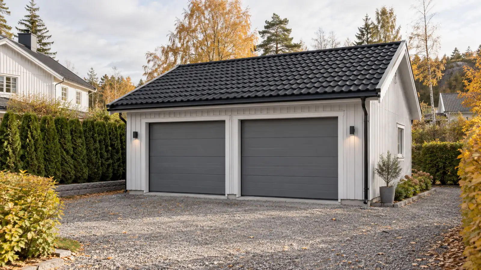 Fristående dubbelgarage med två grå takskjutportar och vit träpanel, grusuppfart och thujahäck i höstljus