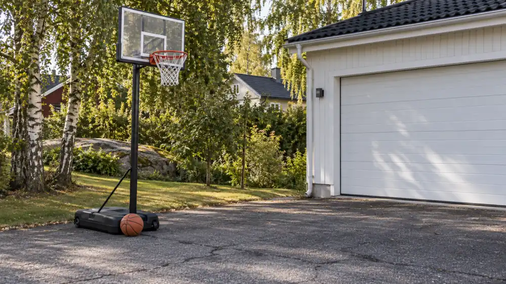 Flyttbar basketkorg med fylld fot på en asfalterad uppfart bredvid en villa med vit garageport och björkar i bakgrunden