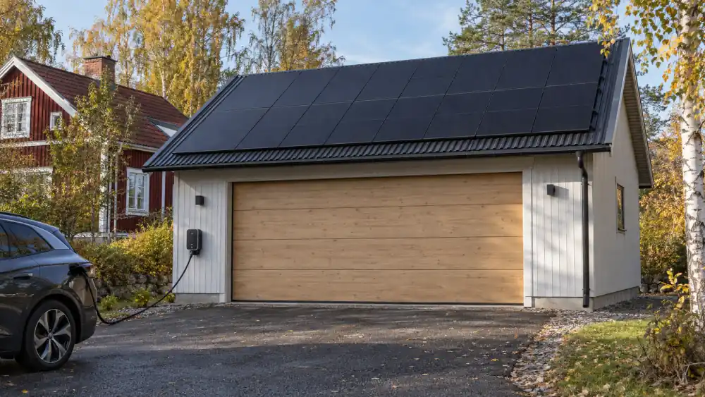 Fristående villagarage med svart plåttak helt täckt av solpaneler, en laddbox på fasaden och en elbil som laddar på uppfarten
